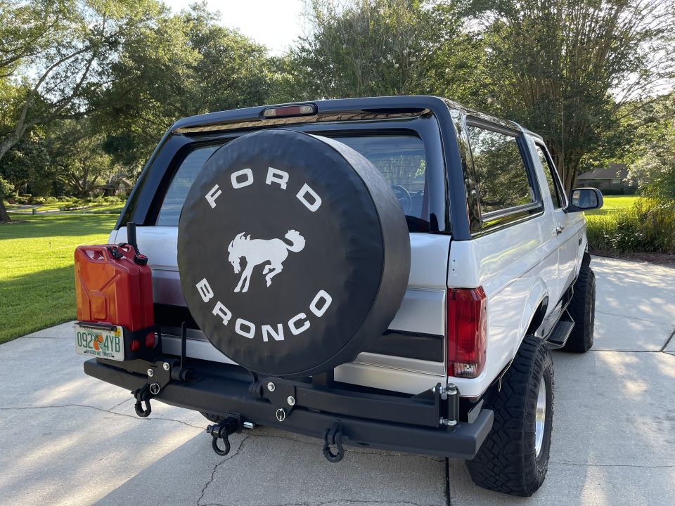 Rear view of a white Ford Bronco with a black spare tire cover showing the Bronco logo and a red gas can mounted on the back