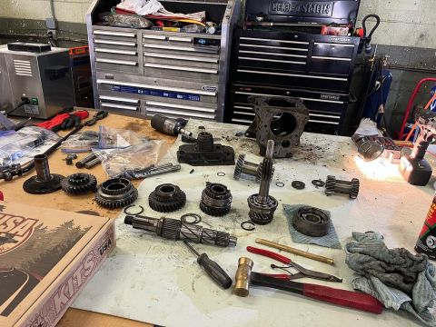 1979 Jeep CJ5 - A workbench is covered in disassembled transmission parts, tools, and a box labeled "KITS USA". A metal cabinet and a parts washer are visi…