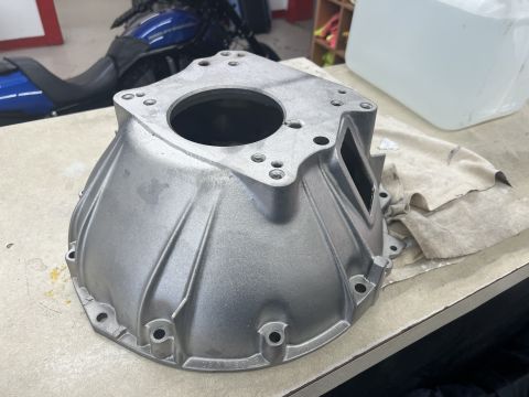 1979 Jeep CJ5 - A silver aluminum bell housing part is on a workbench with a blue motorcycle in the background. The part has a circular opening at the top…