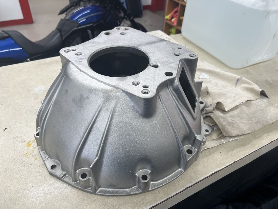 Silver aluminum bell housing part on a workbench, blue motorcycle in background.