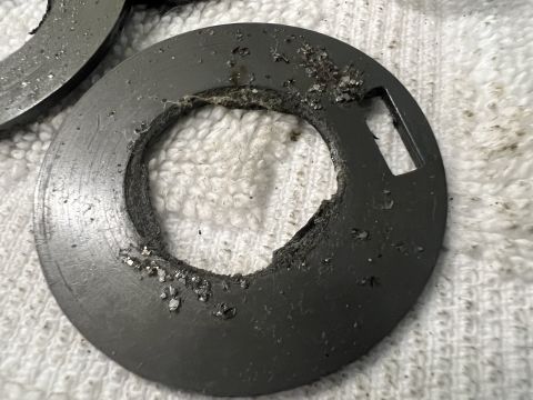 1979 Jeep CJ5 - A close-up view of a damaged, dark-colored metal washer with a rough, torn inner edge. Debris is scattered around the washer and on the whi…