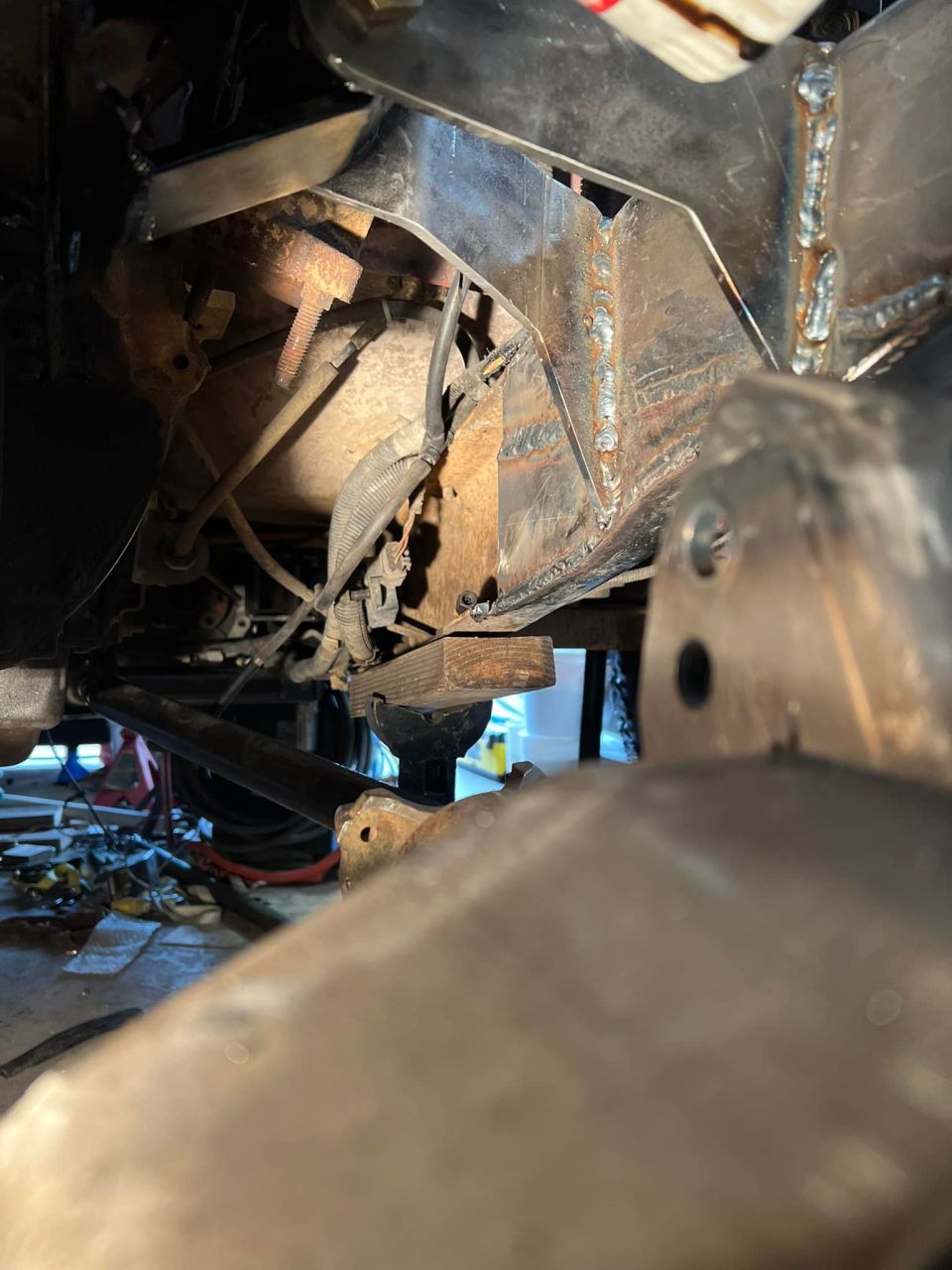 Undercarriage of a vehicle with custom welded metal frame modifications and a wooden support block.