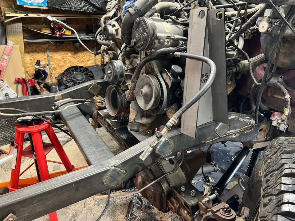 Vehicle frame with engine, custom brackets, and jack stands in a workshop.