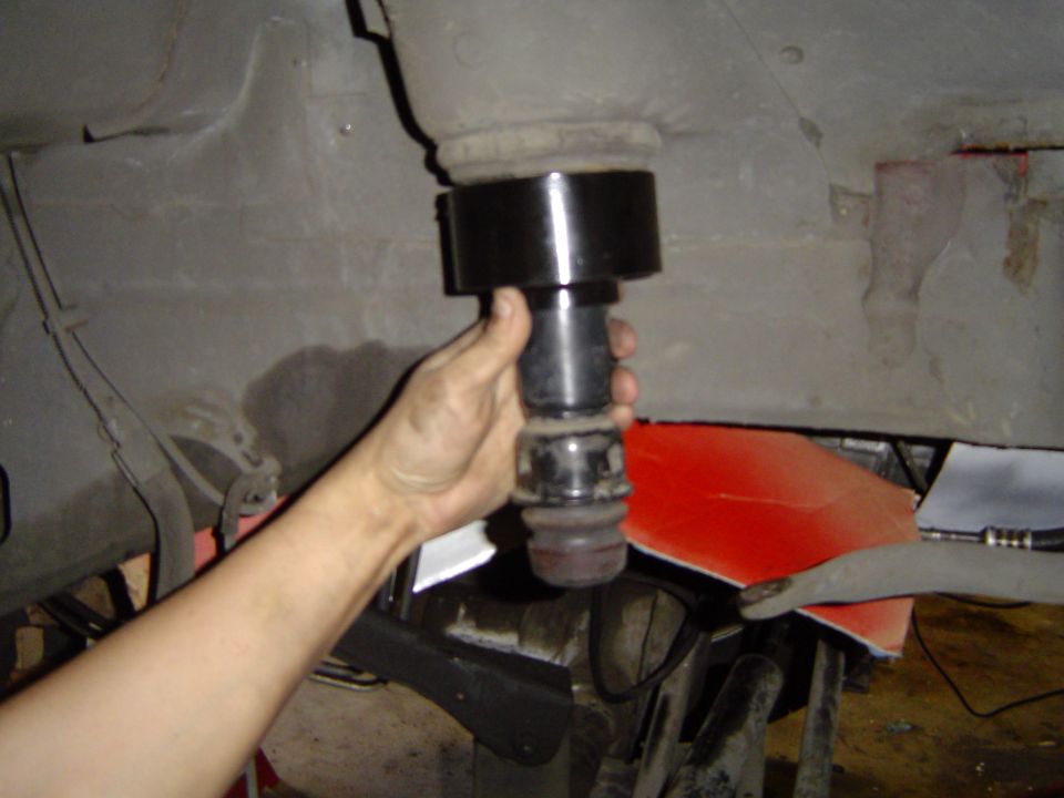 Hand holding a large pressed bushing tool under a vehicle with red panel and frame behind