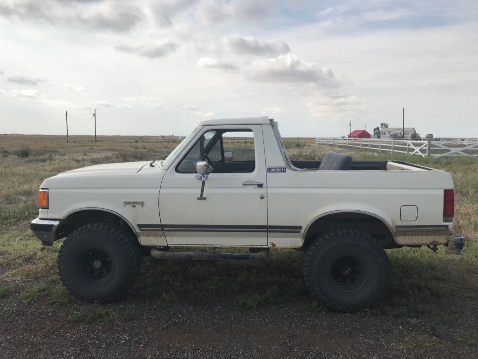 White Ford Bronco parked on dirt with a field and fence in the background