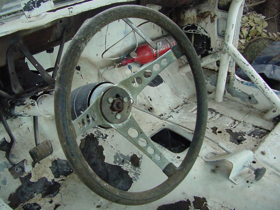 Car interior showing a worn steering wheel, pedals, and a red fire extinguisher mounted on the passenger side