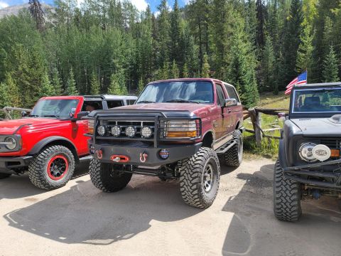 1996 Ford Bronco - Three Ford Broncos are parked outdoors with a wooded mountain backdrop and an American flag.