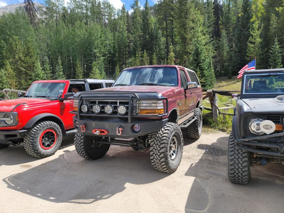 Maroon Ford Bronco with aftermarket bumper and lights, parked next to a red Bronco and a gray Bronco