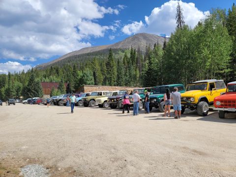 1996 Ford Bronco - A collection of Ford Broncos are parked in a lot with a mountain and trees in the background.