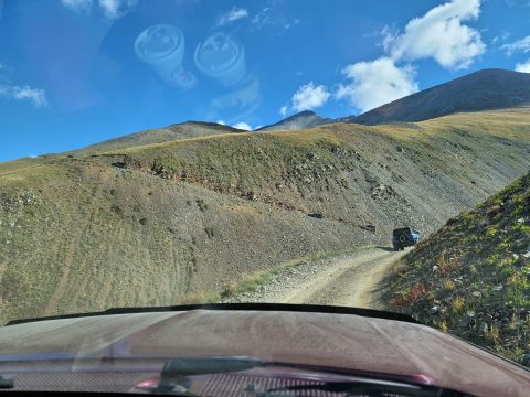 1996 Ford Bronco - Two vehicles navigate a steep dirt road on a mountain under a blue sky with scattered clouds.