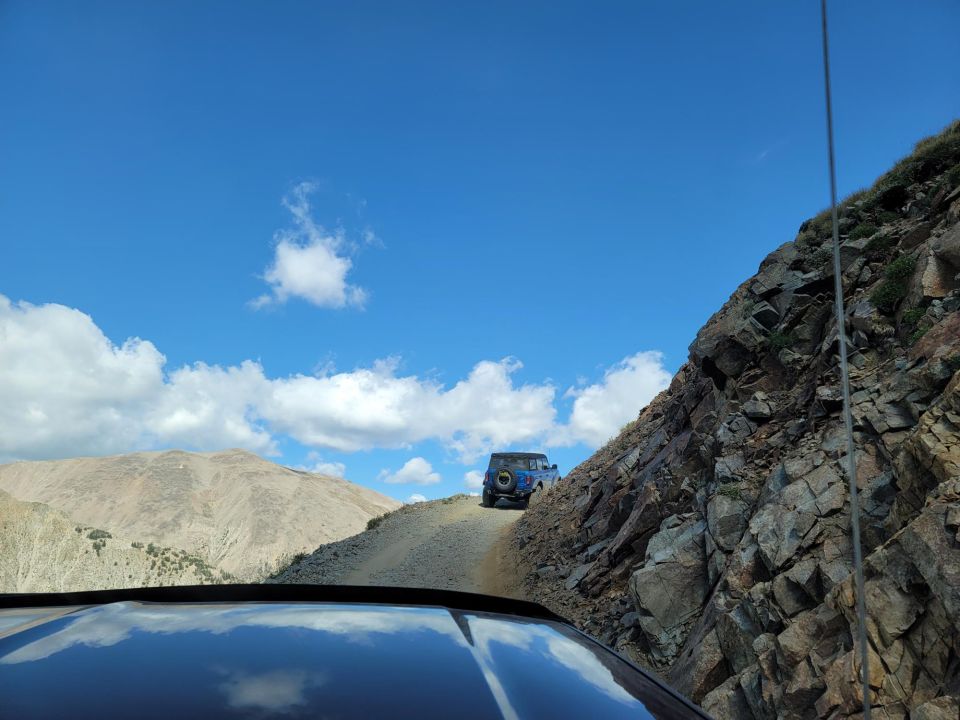 Two blue SUVs drive up a narrow, rocky mountain road under a bright blue sky with scattered clouds