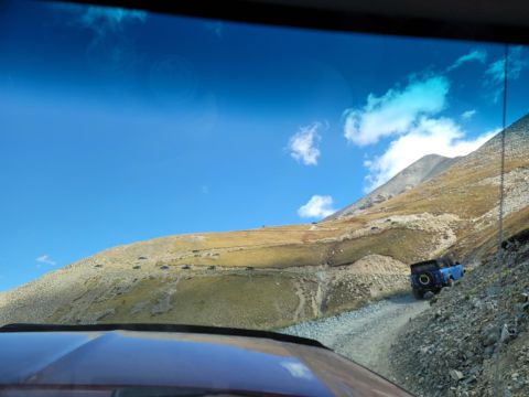 1996 Ford Bronco - A blue SUV navigates a steep, rocky mountain road under a bright blue sky with scattered clouds.