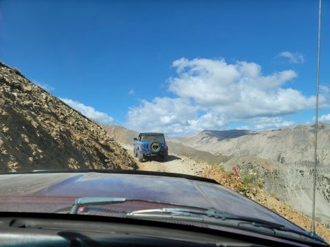 1996 Ford Bronco - Two SUVs navigate a narrow dirt road on a steep mountain pass under a bright blue sky.