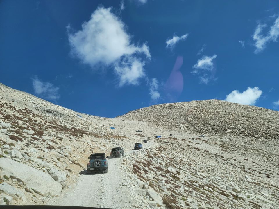 Multiple dark SUVs ascend a gravelly mountain path under a blue sky with scattered clouds