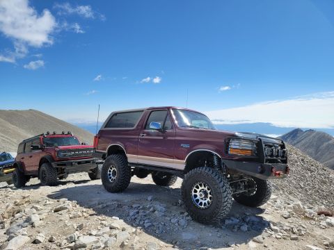 1996 Ford Bronco - Two Ford Broncos, one classic and one modern, ascend a rocky mountain path under a blue sky.