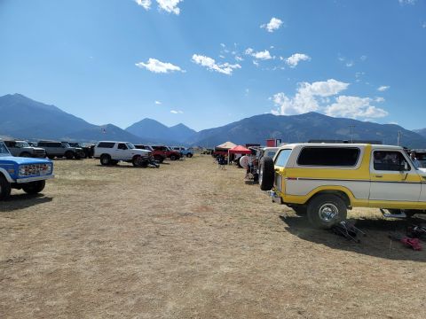 1996 Ford Bronco - A row of vehicles, including several Ford Broncos, are parked outdoors under a partly cloudy sky with mountains in the distance.