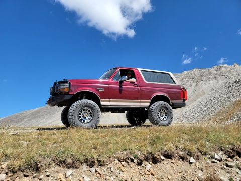 1996 Ford Bronco - A red 1992 Ford Bronco with a tan top is parked on a grassy incline with a mountainous backdrop.