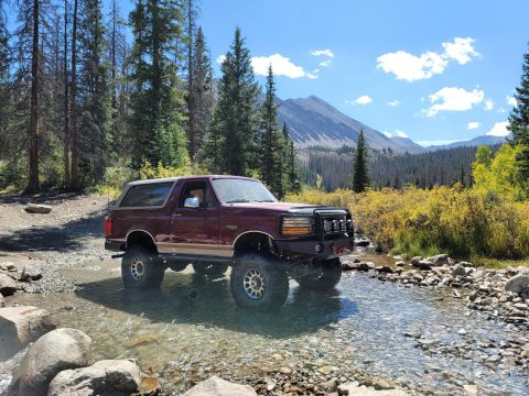 1996 Ford Bronco - A maroon 1996 Ford Bronco navigates a rocky stream bed surrounded by trees and mountains under a partly cloudy sky.