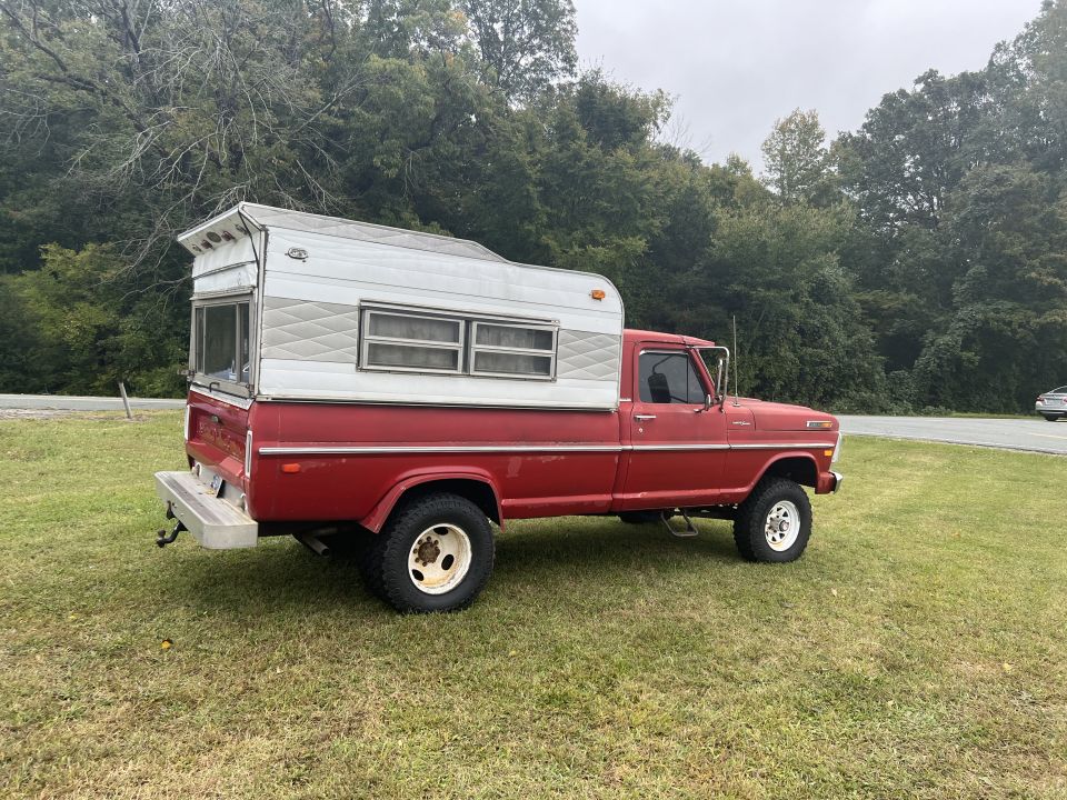 Red 1970 Ford F-250 pickup truck with a white camper shell, parked on grass