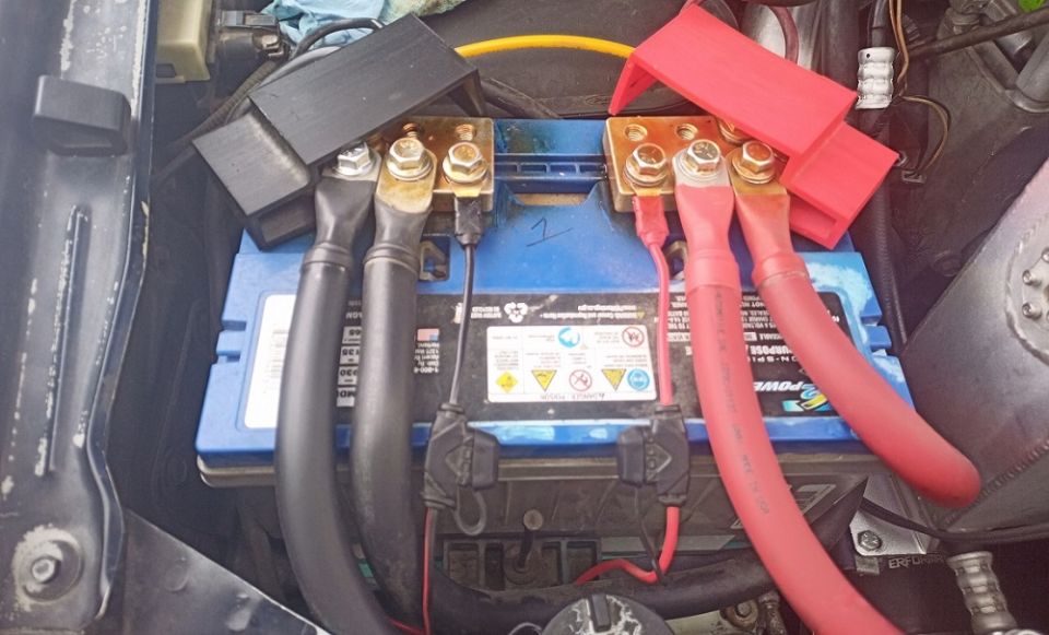 Car battery with red and black cables connected to terminals. Some text and hazard symbols are visible on the battery label