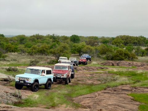 1973 Ford Bronco - A convoy of classic Ford Broncos navigates a , natural landscape under an overcast sky.