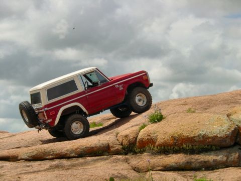 1973 Ford Bronco - A red and white Ford Bronco navigates a steep, rocky terrain under a cloudy sky.
