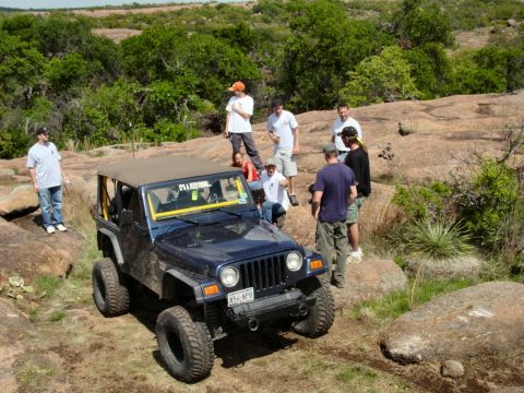 1973 Ford Bronco - A blue Jeep Wrangler navigates a rocky trail with several onlookers gathered around.