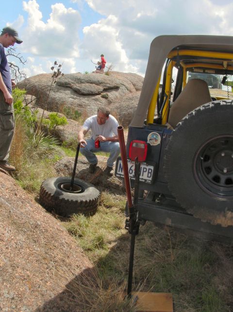 1973 Ford Bronco - Off-roading group changes a tire on a Jeep Wrangler in a rocky, outdoor landscape.