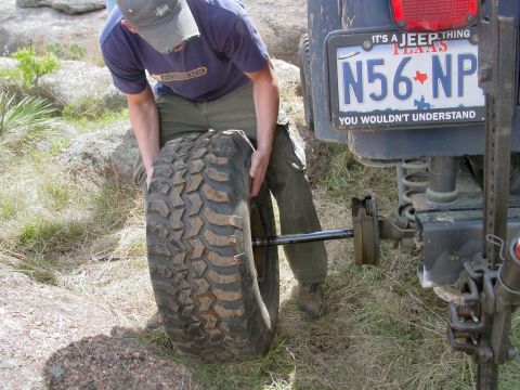 1973 Ford Bronco - A person is changing a tire on a Jeep, which is parked on rocky terrain.
