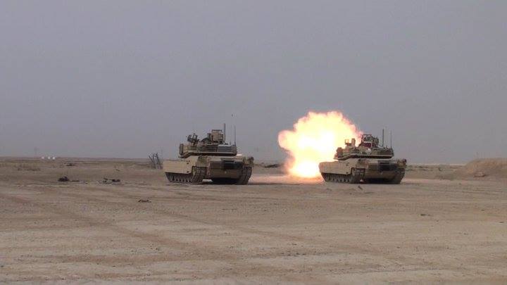 Two M1 Abrams tanks in a desert, one firing with a large explosion.