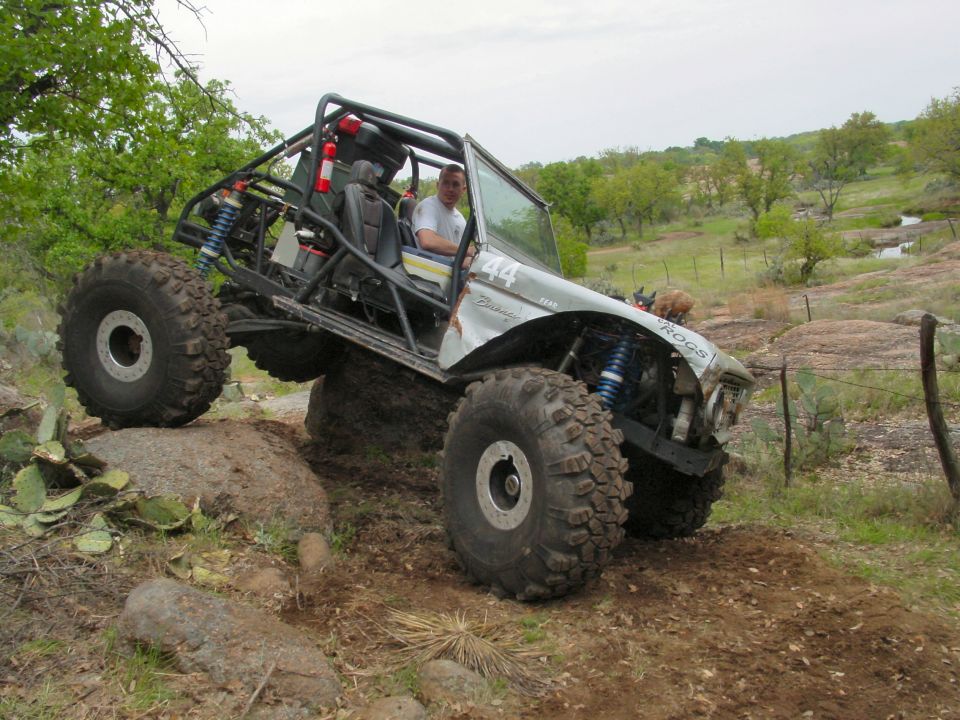 Silver 1973 Ford Bronco modified for off-roading, climbing a rock with large tires and a roll cage