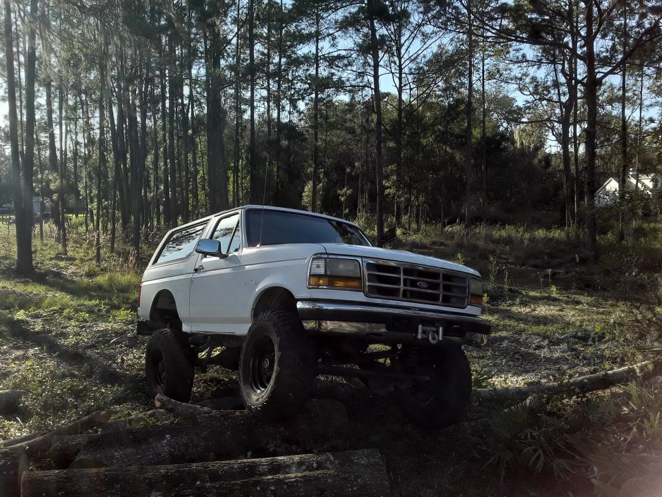 White 1994 Ford Bronco parked on a dirt path in a forest.