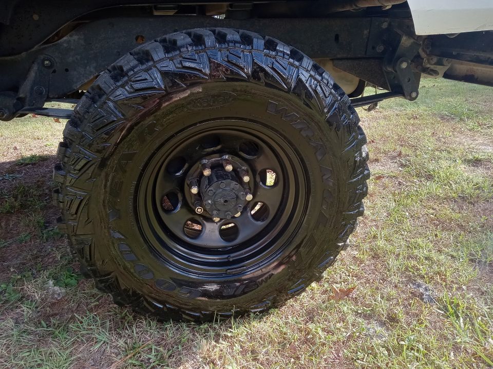 Close-up of a black off-road tire and wheel on a vehicle. Text on tire is illegible