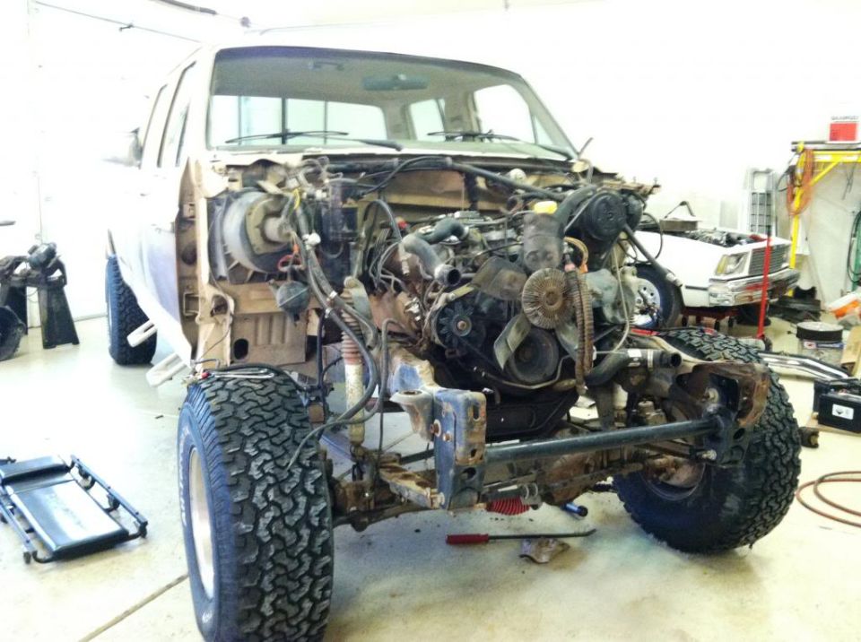 Truck engine bay with engine removed, showing chassis and front suspension. Another vehicle visible in background