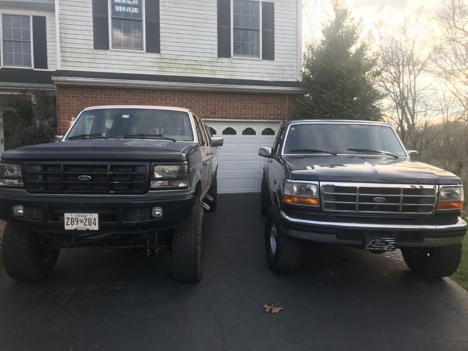 Two dark-colored Ford trucks parked in front of a house and garage. The truck on the left has a lifted suspension and larger tires