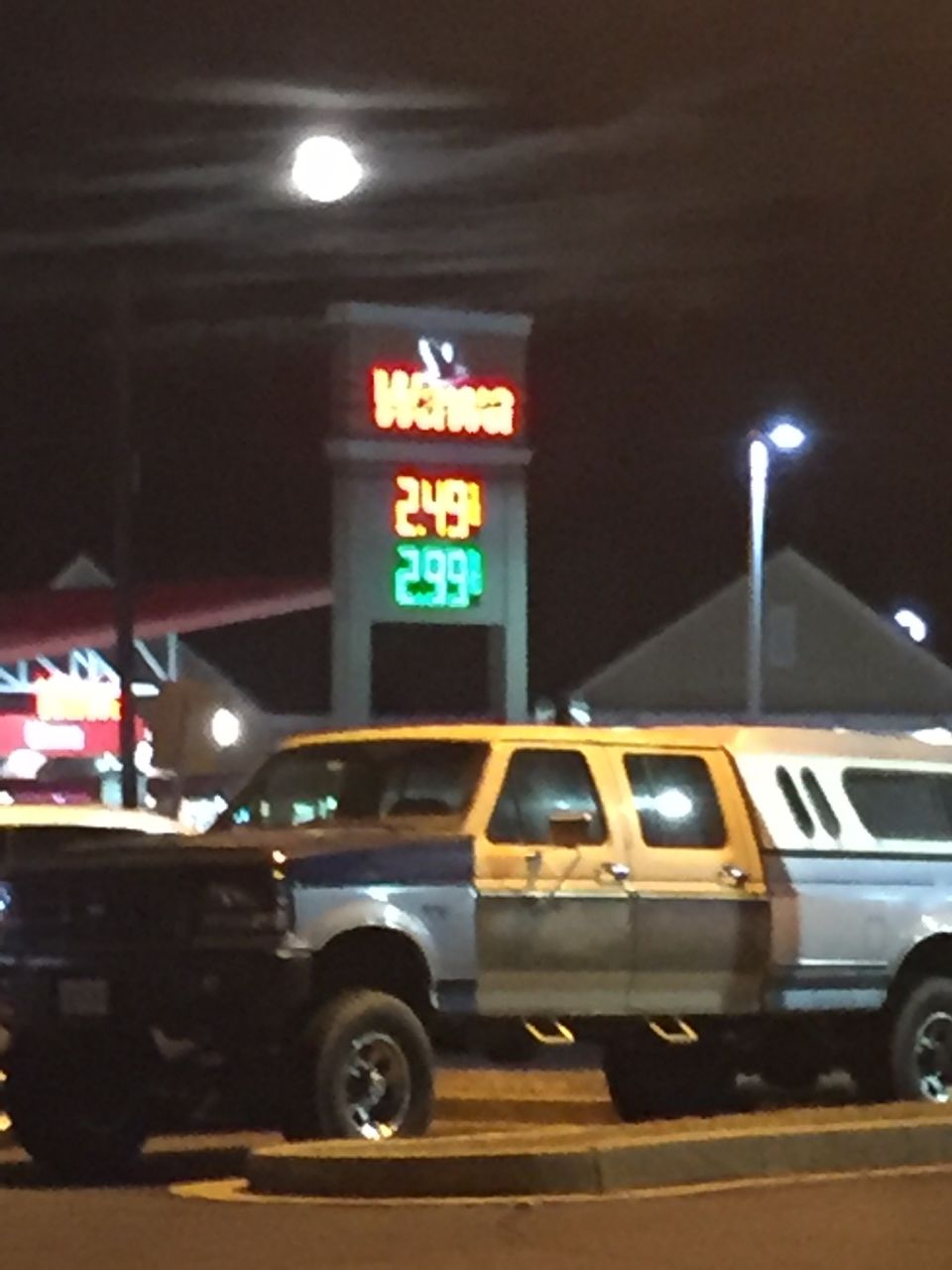 Lifted pickup truck with camper shell parked at Wawa gas station at night.