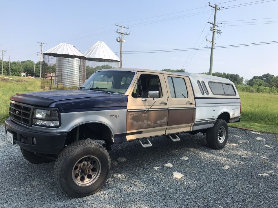 Modified blue and brown crew cab pickup truck with a white camper shell parked on gravel.
