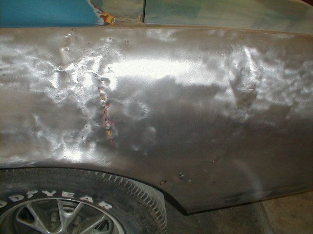 Car's rear quarter panel is bare metal, showing dents and weld repairs. A Goodyear tire is visible