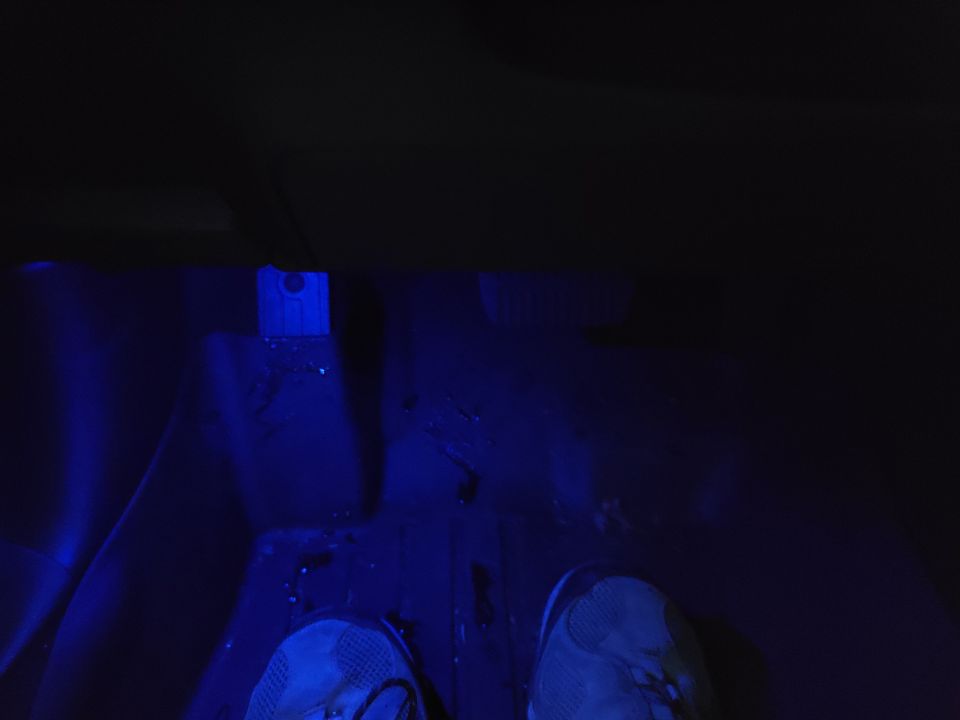Interior of a vehicle with blue ambient lighting, showing pedals and feet on the floor.