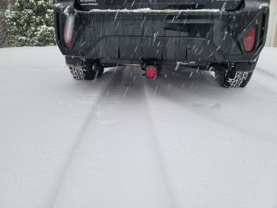 Rear view of a dark Subaru Crosstrek in the snow with tire tracks and a trailer hitch light.