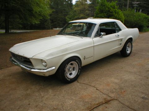 1967 Ford Mustang - A white 1967 Ford Mustang coupe is parked on a concrete driveway, with trees in the background.