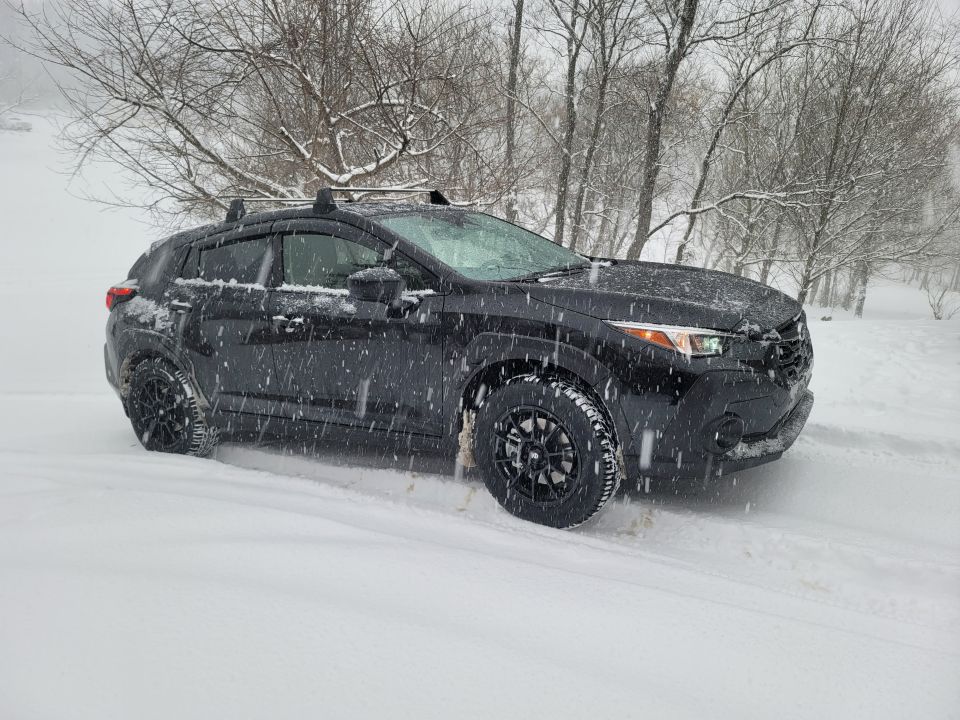 Black Subaru Crosstrek in a snowstorm with roof racks and aftermarket wheels.