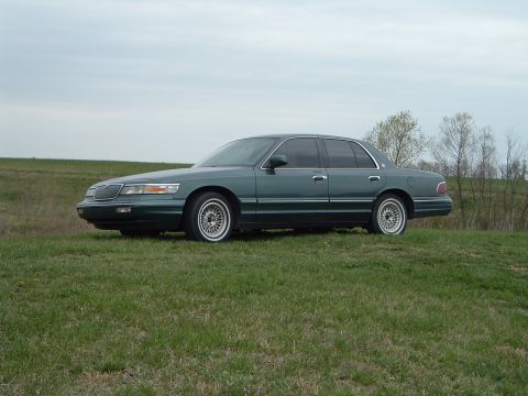 1995 Mercury Grand Marquis - A green 1995 Mercury Grand Marquis is shown from the side, parked on a grassy incline undercast sky.