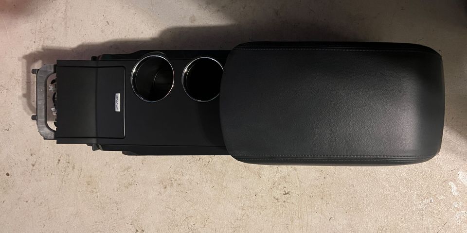 Black center console with two chrome cup holders and leather armrest lid, removed and lying on concrete floor