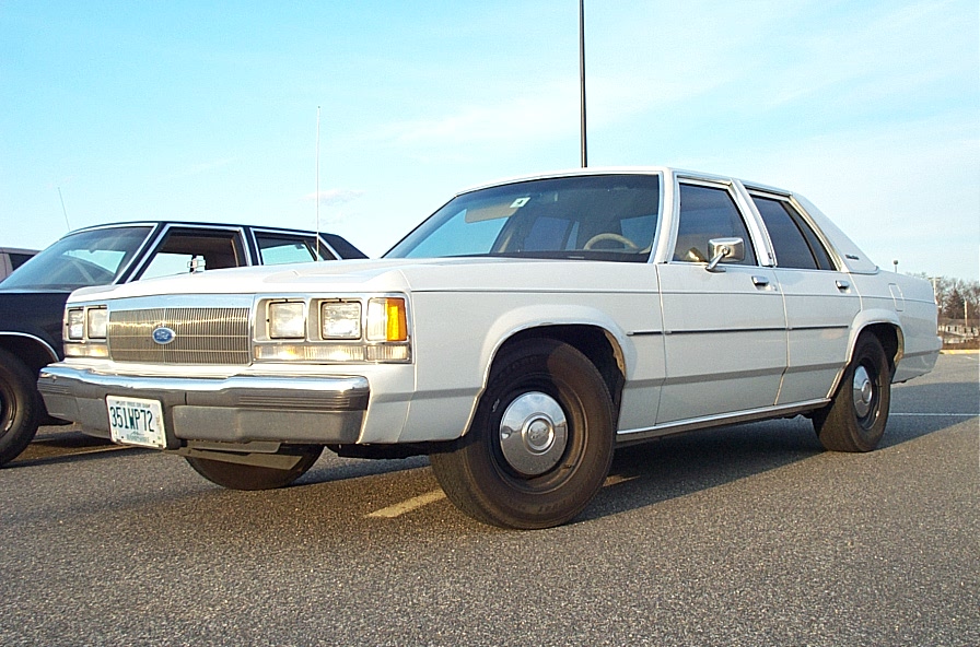 White 1980s Ford sedan parked next to a dark sedan, front quarter view