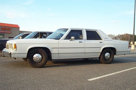 1991 Ford LTD Crown Victoria - A white sedan is parked in a lot next to another vehicle, with a building visible in the background.