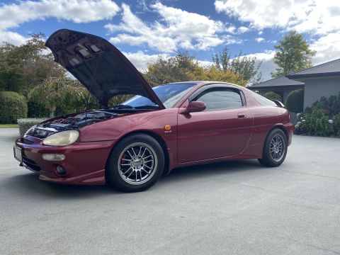 1993 Mazda MX-3 - A red 1993 Mazda MX-3 coupe is parked outside a house with its hood open.