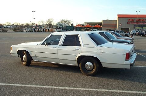 1991 Ford LTD Crown Victoria - A white sedan is parked in a lot with other vehicles and a store in the background.