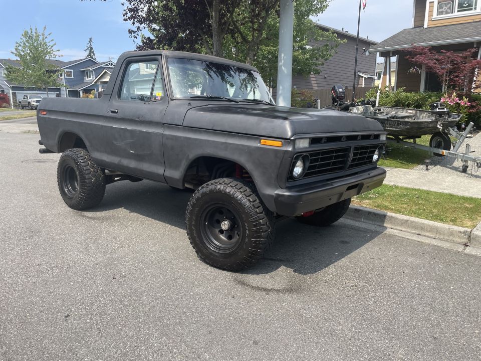 Lifted matte black 1970s Ford Bronco with off-road tires parked on street, boat on trailer in background.