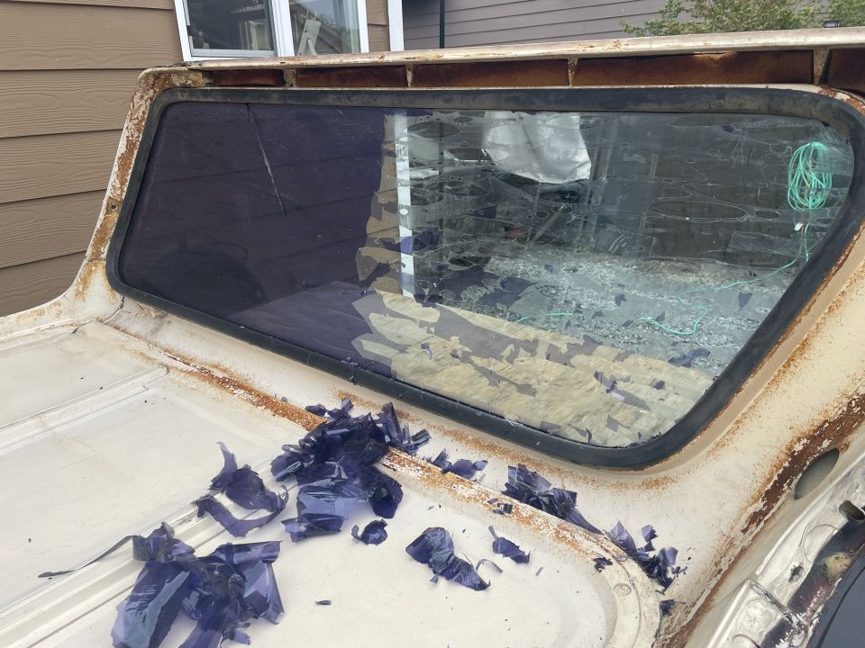 Damaged rear window of a light-colored vintage SUV with shattered purple glass and rust.